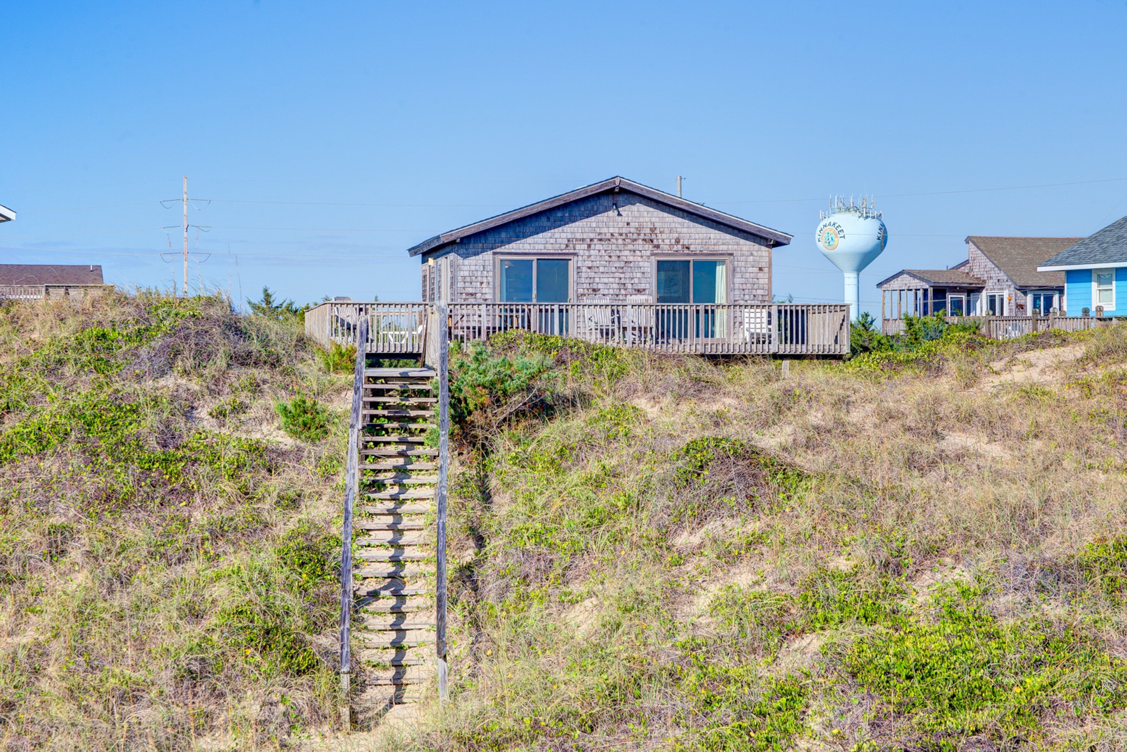 A coastal building with a wooden staircase leading to a porch, surrounded by vegetation and a clear sky.