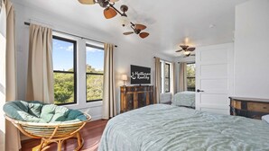 A bedroom with a large bed, a wicker chair, a ceiling fan, and a view of greenery outside. - Perfect Retreat with Private Hot Tub, Panoramic Views, Full Kitchen pet-friendly (Buxton)
