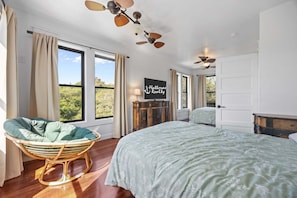 A bedroom with a large bed, a wicker chair, a ceiling fan, and a view of greenery outside.