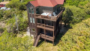 A two-story wooden cabin with a balcony, surrounded by dense greenery and overlooking a body of water. - Perfect Retreat with Private Hot Tub, Panoramic Views, Full Kitchen pet-friendly (Buxton)