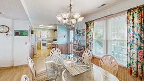 A dining area with a glass table, white chairs, and a chandelier. There is a kitchen in the background with white cabinets and a microwave. - Coastal home with large deck, close to beach and local attractions (Holden Beach)