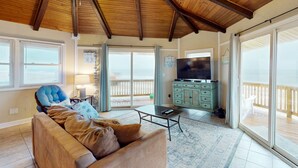 A spacious living room with a wooden ceiling, a large television, a sofa, and a sliding glass door leading to a balcony. - Dog-friendly beachfront home with beach access, ocean views (North Topsail Beach)