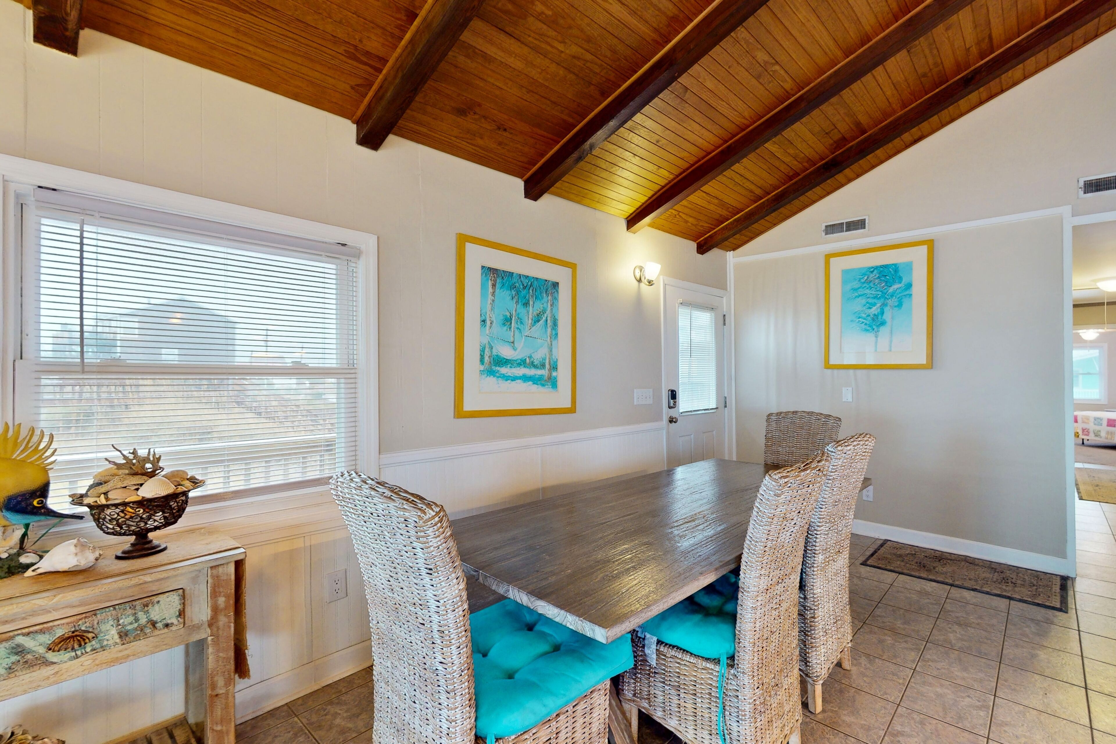 A dining area with a wooden table and wicker chairs, framed artwork on the walls, and a window with blinds.