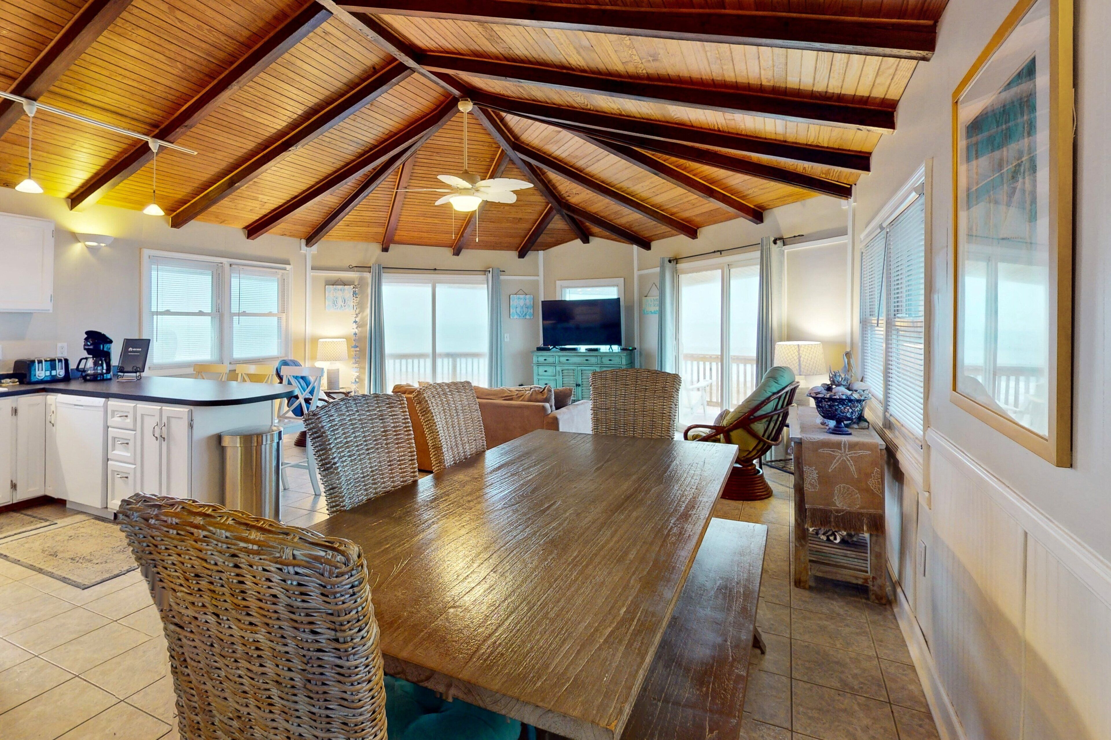 A spacious kitchen with a wooden ceiling, a dining area with wicker chairs, and a large TV in the background.