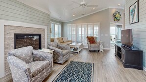 A spacious living room with a fireplace, a ceiling fan, and a large window. - Dog-friendly marsh view home with large deck, close to beach access (Holden Beach)