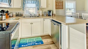 A kitchen with white cabinets, a stainless steel range hood, a dishwasher, and a blue and green beach-themed mat on the floor.