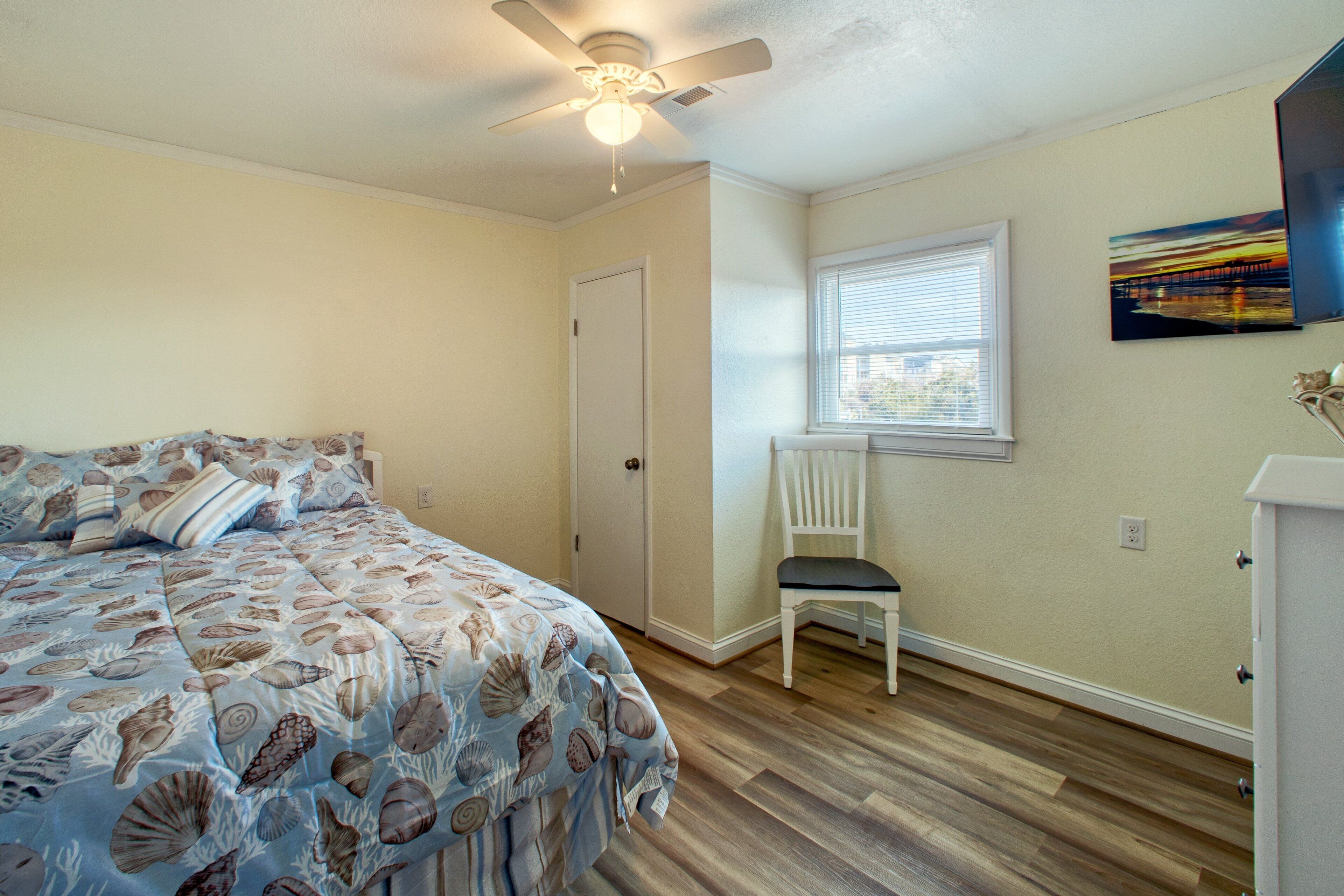 A bedroom with a bed, a chair, a ceiling fan, a window, and a painting on the wall.