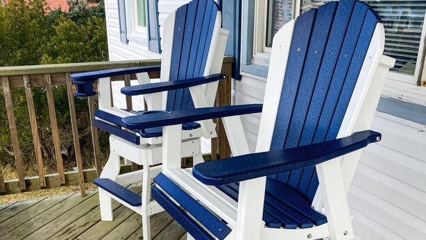 Two Adirondack chairs on a wooden deck with a white house in the background.