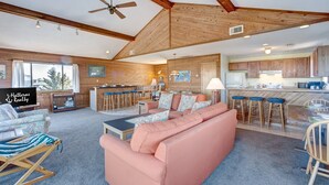 A spacious living area with a large sofa, a coffee table, and a television. There is a kitchen area with bar stools and a dining table. The room has wooden beams on the ceiling and a large window providing natural light. - Brigadune 143: Dog Friendly, Ocean view, 3 lots from beach (Avon)