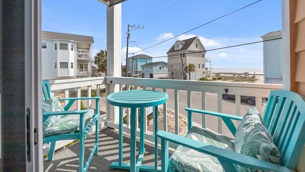A balcony with turquoise chairs and a small table, overlooking a beach and residential buildings.