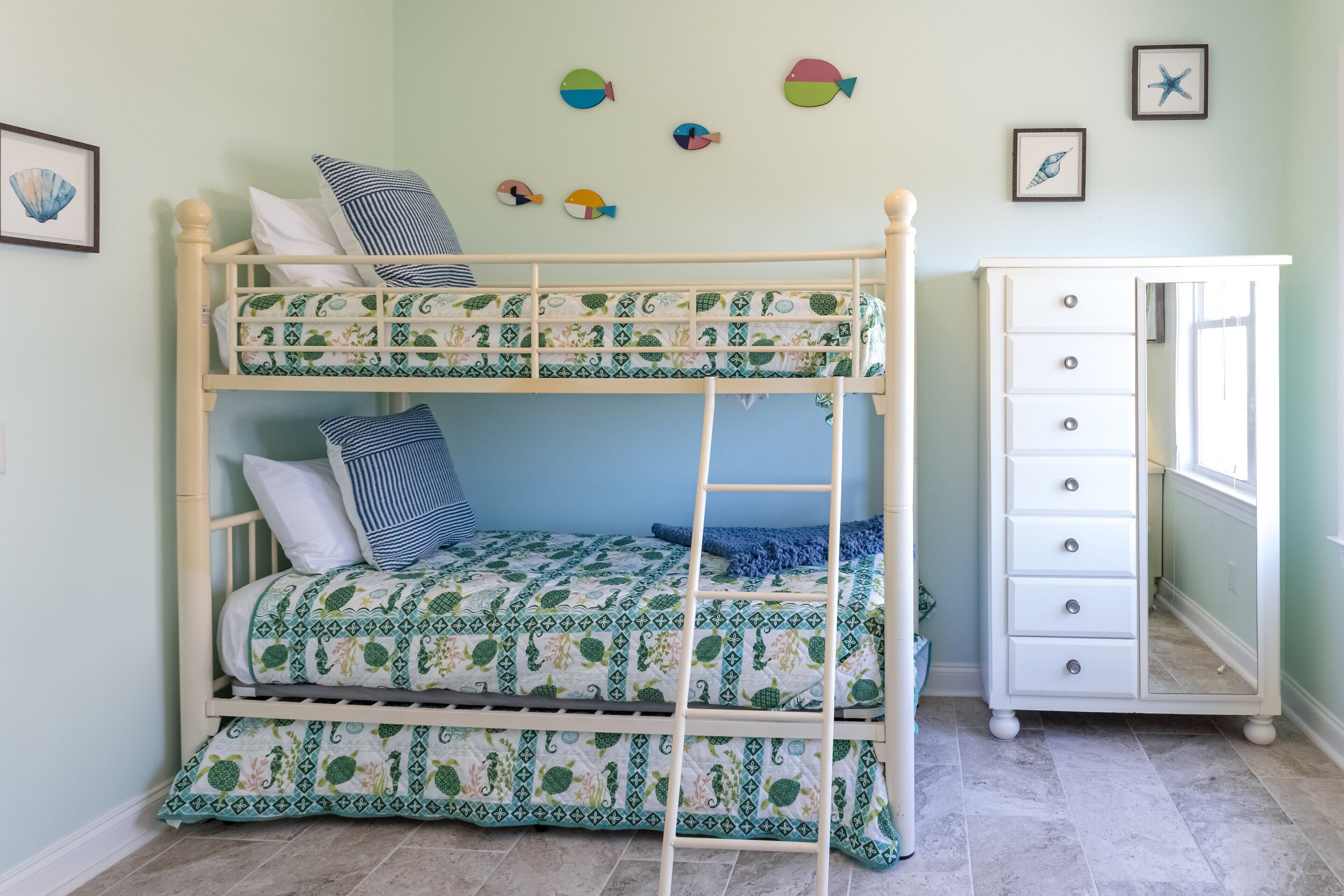 Una cama litera con colcha estampada, un aparador con cajones y decoraciones en la pared con motivos de peces y estrellas de mar.