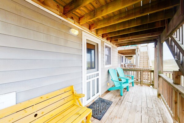 A wooden deck with a bench and Adirondack chairs, a covered area with a staircase leading to an upper level.