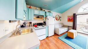 Une cuisine compacte avec des meubles de couleur turquoise, un poêle blanc et un réfrigérateur blanc. On y trouve également un canapé blanc et un petit tabouret en bois dans l’espace de vie adjacent.