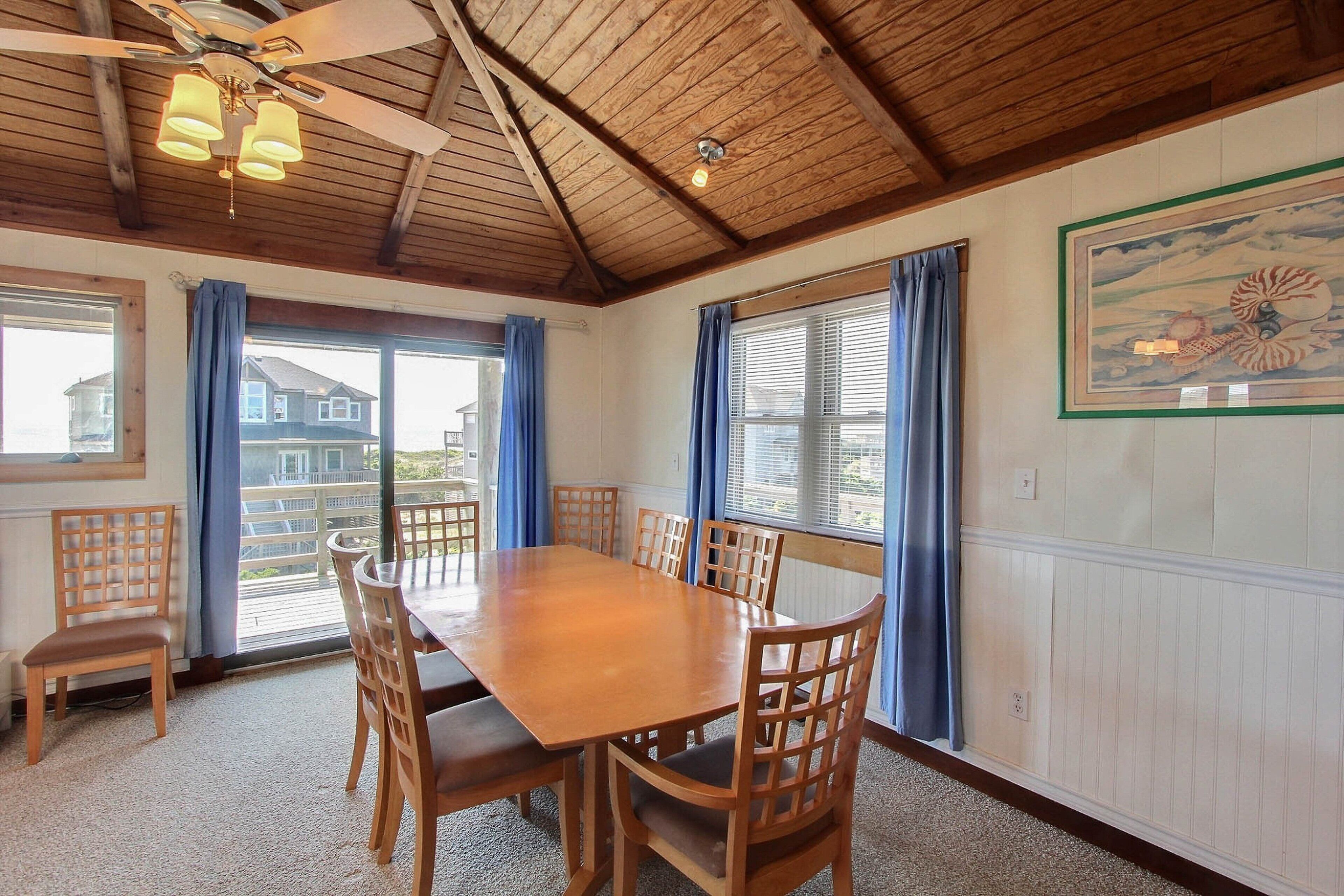 A dining area with a wooden table and chairs, large windows with blue curtains, and a ceiling fan.
