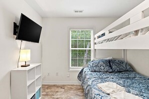 A bedroom with a bed, a TV mounted on the wall, a window with blinds, and a shelf unit. - Seas the Day | Screened Porch & Bikes | Walk to Beach & Intracoastal Waterway (Oak Island)