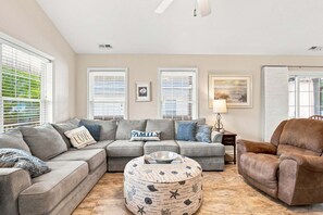 A living room with a sectional sofa, ottoman, and armchair, a side table with a lamp, and artwork on the walls. - Seas the Day | Screened Porch & Bikes | Walk to Beach & Intracoastal Waterway (Oak Island)