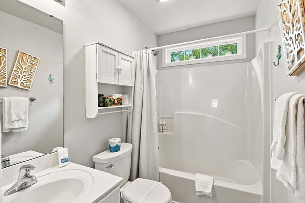 A bathroom with a sink, toilet, and shower. There is a window above the shower, and a towel rack is visible. - Seas the Day | Screened Porch & Bikes | Walk to Beach & Intracoastal Waterway (Oak Island)