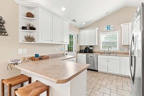 A modern kitchen with white cabinets, a large island with bar stools, a dishwasher, and a refrigerator. - Seas the Day | Screened Porch & Bikes | Walk to Beach & Intracoastal Waterway (Oak Island)