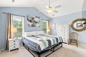 Une chambre à coucher comprenant un lit, une table de chevet, une commode, une chaise, un ventilateur de plafond et des œuvres d’art accrochées aux murs.