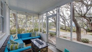 A covered porch with a blue sofa, a stone fireplace, and large windows overlooking a street. - My Three Buoys: Cozy stay close to everything with backyard, bikes, grill, & washer/dryer (Oak Island)