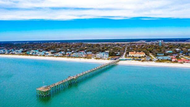 An aerial view of a coastal area with a pier extending into the water, surrounded by residential buildings and a beach. - My Three Buoys: Cozy stay close to everything with backyard, bikes, grill, & washer/dryer (Oak Island)