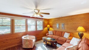A cozy living room with wooden walls, a ceiling fan, a sofa, a coffee table, and two lamps. - Waterfront House w/ Water View, Shared Pool, Private Washer/Dryer, & Free WiFi (Frisco)