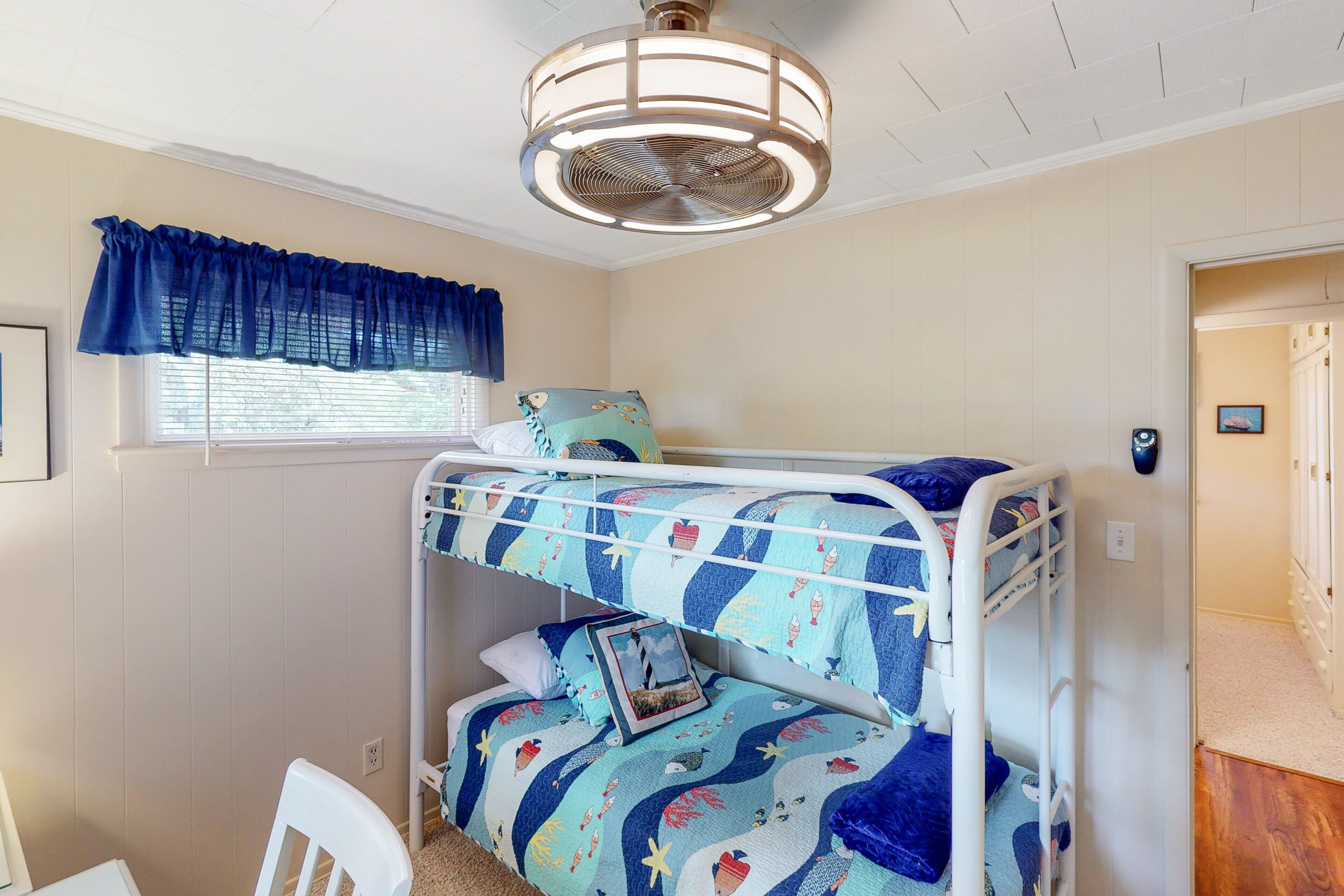 A bunk bed room with a ceiling fan, a window with blue curtains, and a framed picture on the bed.