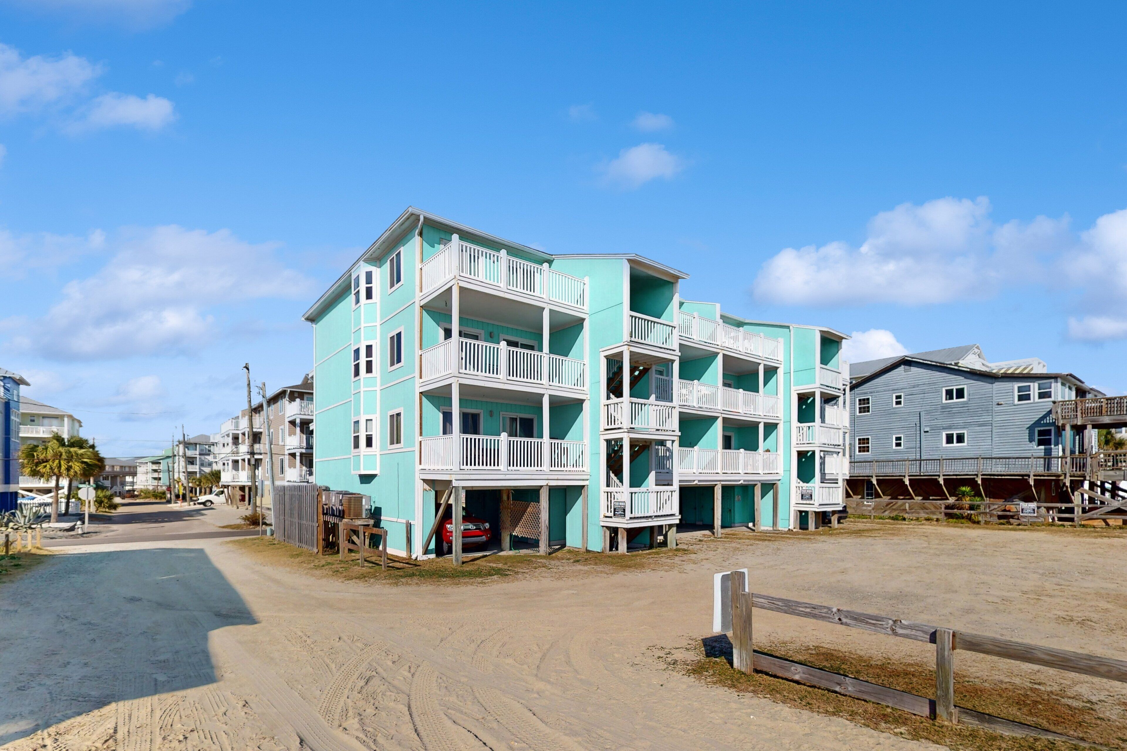 Um edifício de vários andares com varandas, chão de areia e céu claro.
