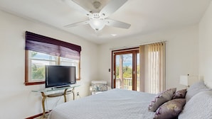 Une chambre à coucher avec un lit, un téléviseur posé sur une table en verre, un ventilateur de plafond et une vue à travers une porte.