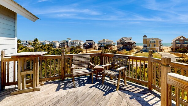 Eine Holzterrasse mit zwei Stühlen und einem Tisch, mit Blick auf einen Strand und eine Reihung von Häusern.