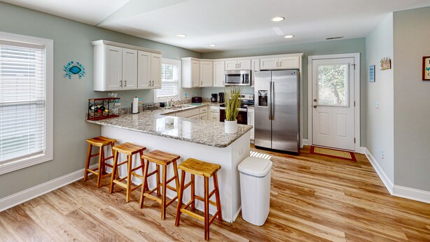 A modern kitchen with white cabinets, a granite island with bar stools, stainless steel appliances, and a wooden floor. - Oak Island Oasis: Updated, dog-friendly home near the beach with fenced yard (Oak Island)