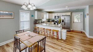 A modern kitchen with white cabinets, a stainless steel refrigerator, and a dining area with wooden chairs and a table. - Oak Island Oasis: Updated, dog-friendly home near the beach with fenced yard (Oak Island)
