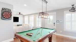 Ein Billardtisch mit Kugeln und Spielstangen, eine Dartscheibe, ein Sofa und ein Deckenventilator in einem Raum mit großen Fenstern und Holzböden.