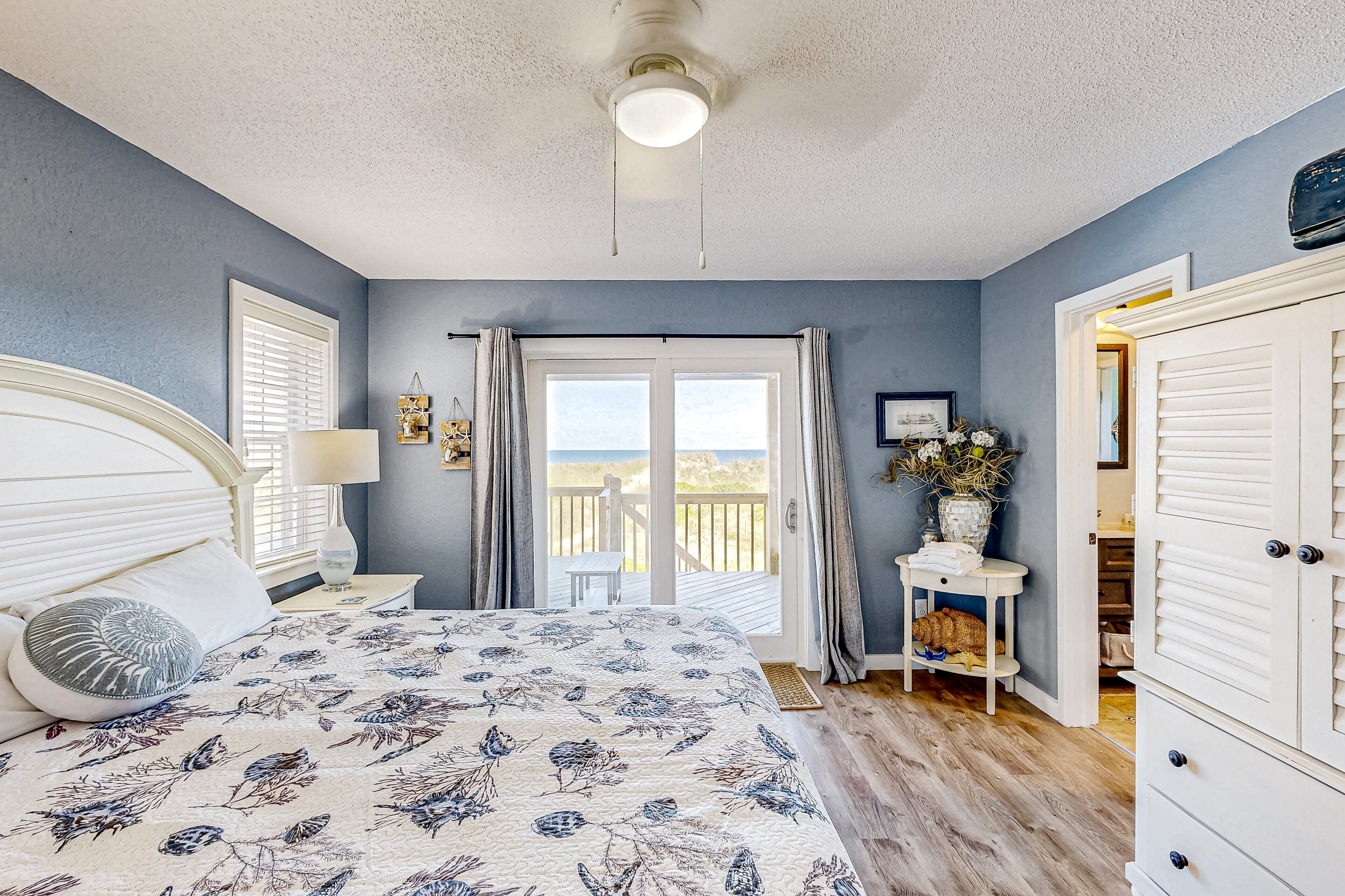 A bedroom with a bed, a nightstand, a window with curtains, a door, and a view of the outdoors.