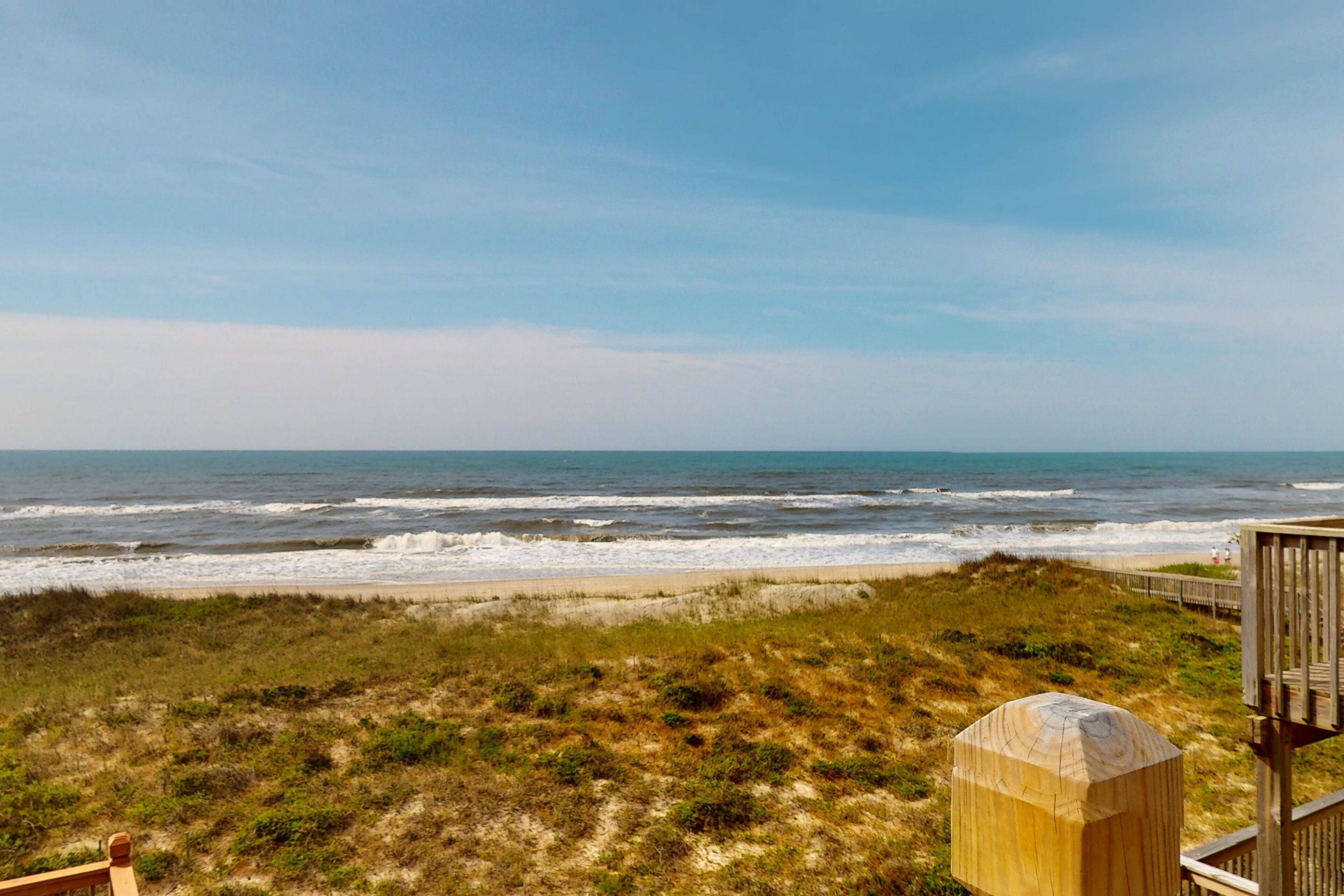 Vista de la playa con una terraza de madera y una duna.