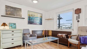 Un salon avec une table basse en verre, un canapé, un fauteuil, une commode et des œuvres d’art accrochées aux murs.