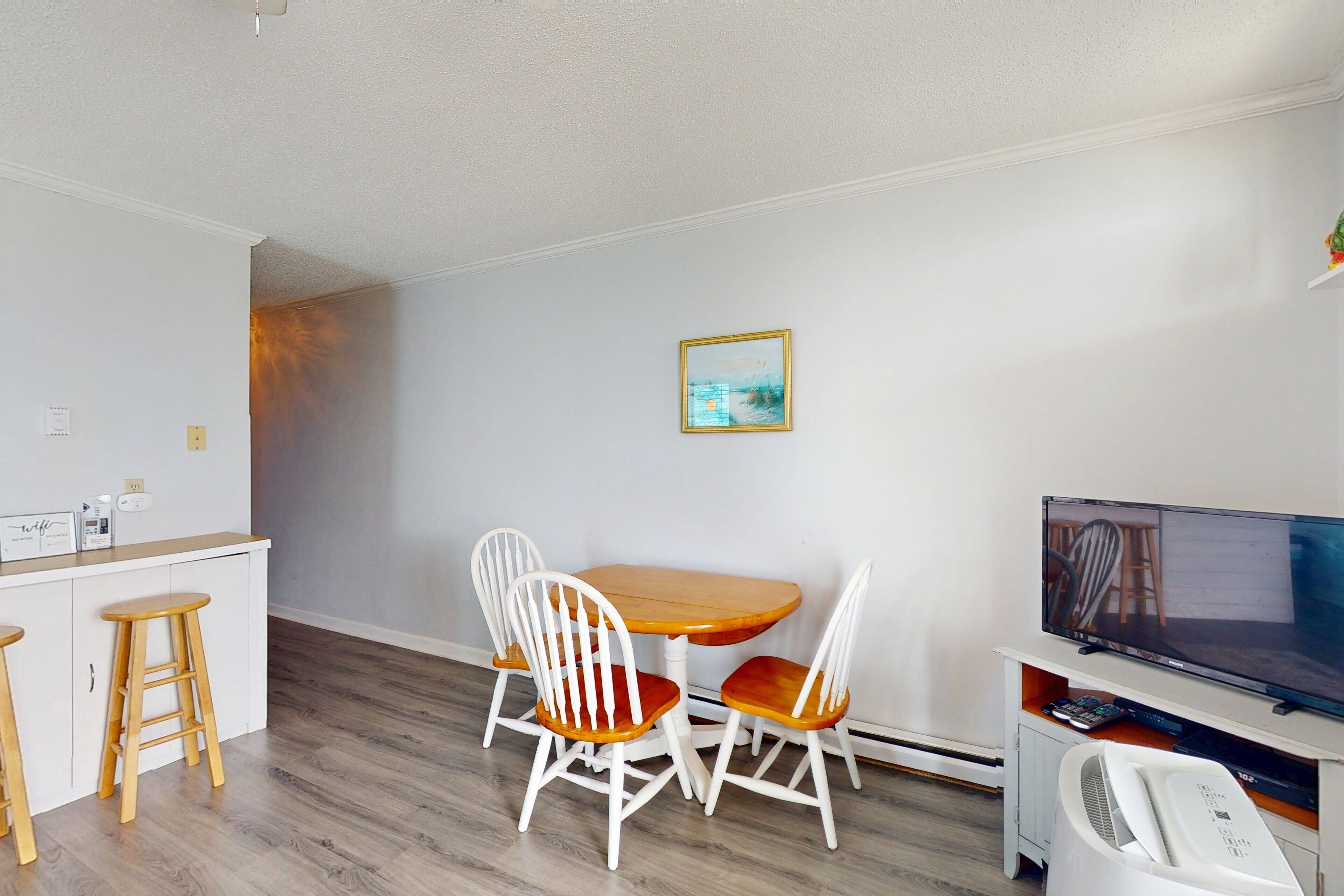 A small, clean room with a wooden dining table and four white chairs, a TV, and a small white fan.