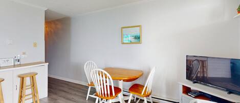 A small, clean room with a wooden dining table and four white chairs, a TV, and a small white fan.