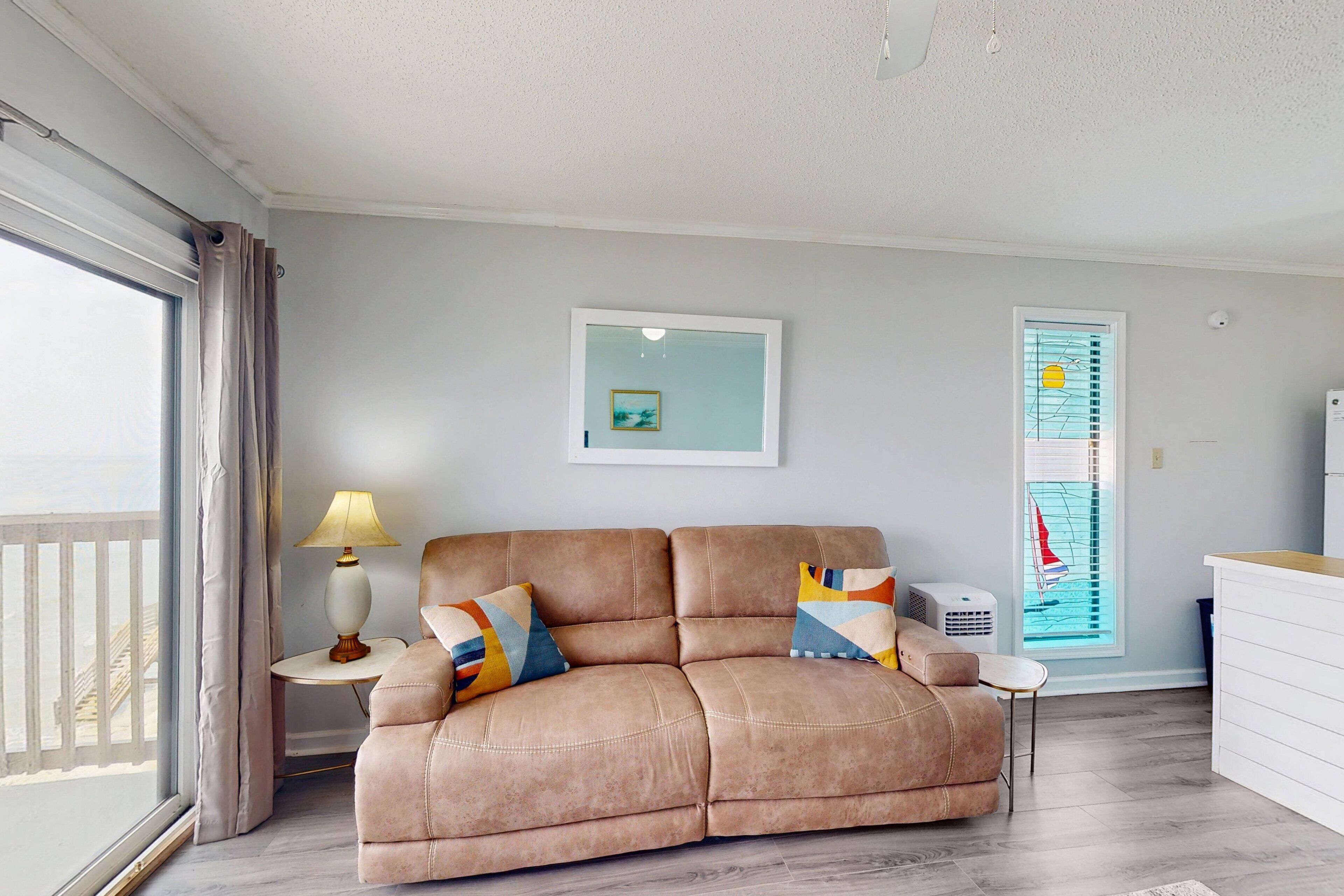A living room with a beige sofa, a small table with a lamp, a framed picture on the wall, and a window with a view of a balcony.