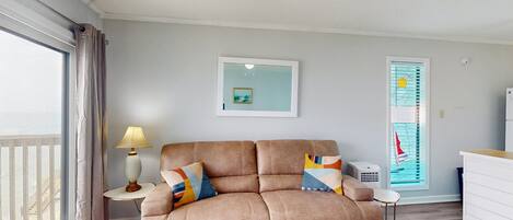 A living room with a beige sofa, a small table with a lamp, a framed picture on the wall, and a window with a view of a balcony.