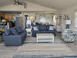 A living room with a sofa, coffee table, and patterned chair. There is a kitchen in the background with cabinets and a refrigerator. - Dreamy dog-friendly getaway with ocean views, beach access, fast WiFi, and deck (Frisco)