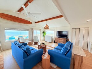A living room with two blue sofas, a TV, a ceiling fan, and a view of the ocean.