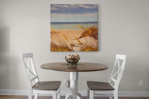 A dining table with two chairs, a painting of a beach scene, and a bowl on the table.