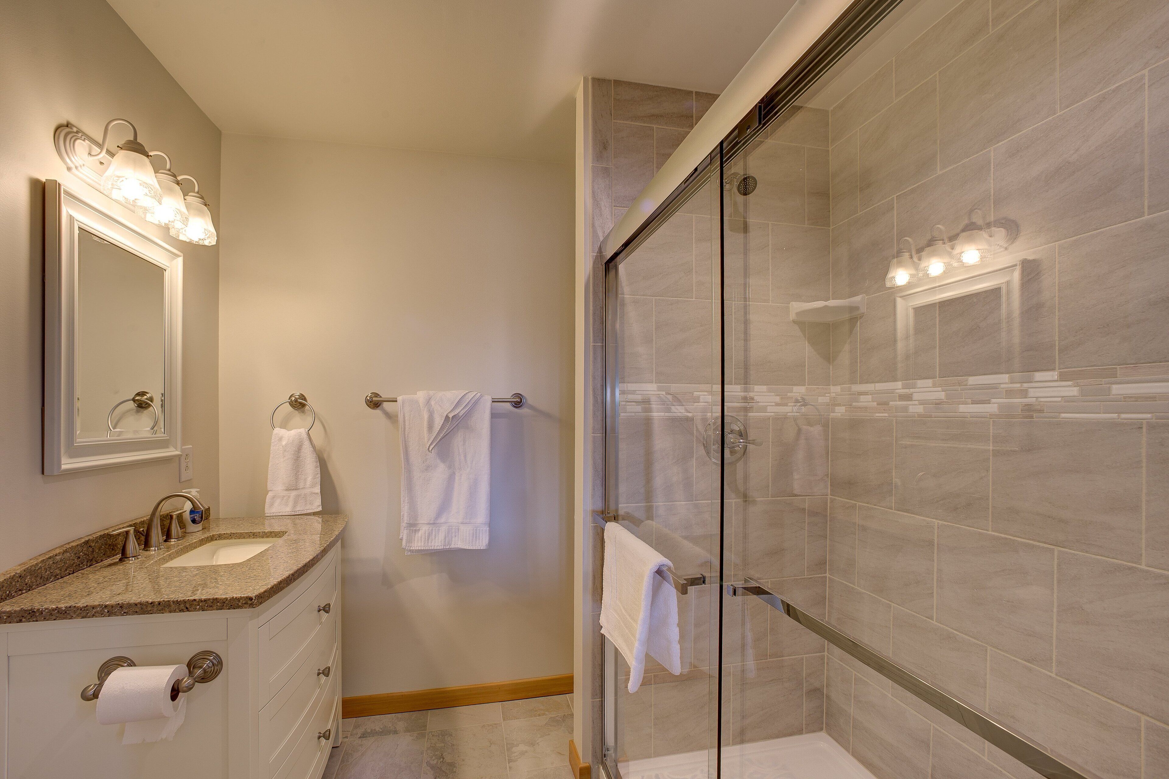 A bathroom with a granite countertop sink, a mirror with lights, a glass shower enclosure, and beige tiled walls.