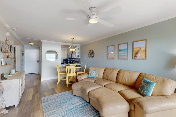 A living room with a beige sectional sofa, a dining area with a wooden table and chairs, and a kitchen in the background. - Pelican's Penthouse:  Snowbird friendly top-floor condo with ocean view balcony (Caswell Beach)