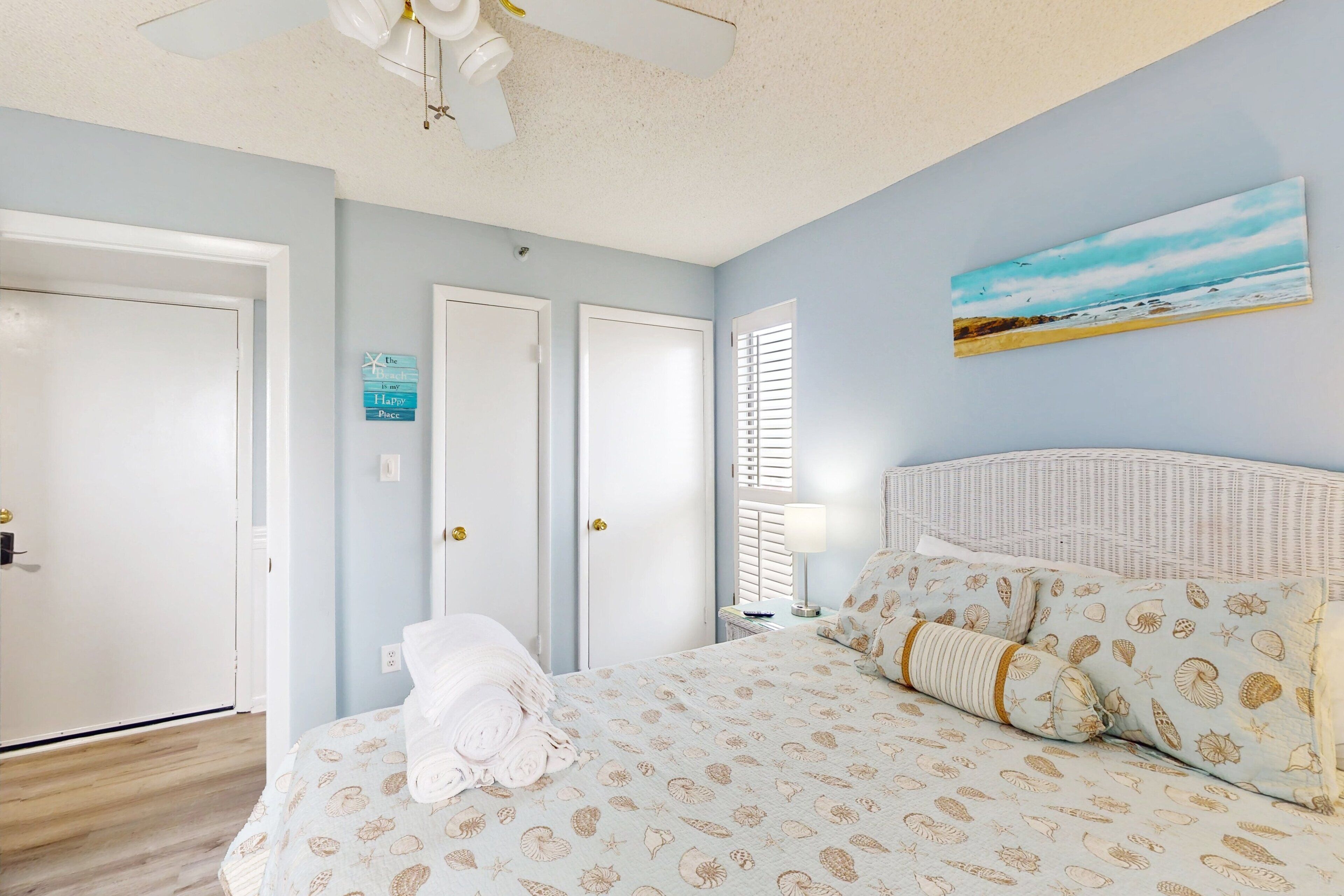 A bedroom with a bed, a ceiling fan, a painting of a beach, and a sign on the wall.