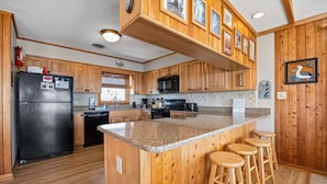 Eine Küche mit Holzausstattung, einer Granit Arbeitsplatte, einem schwarzen Kühlschrank, einer Mikrowelle und einem Spülbecken. An der Kochinsel stehen drei hölzerne Hocker.