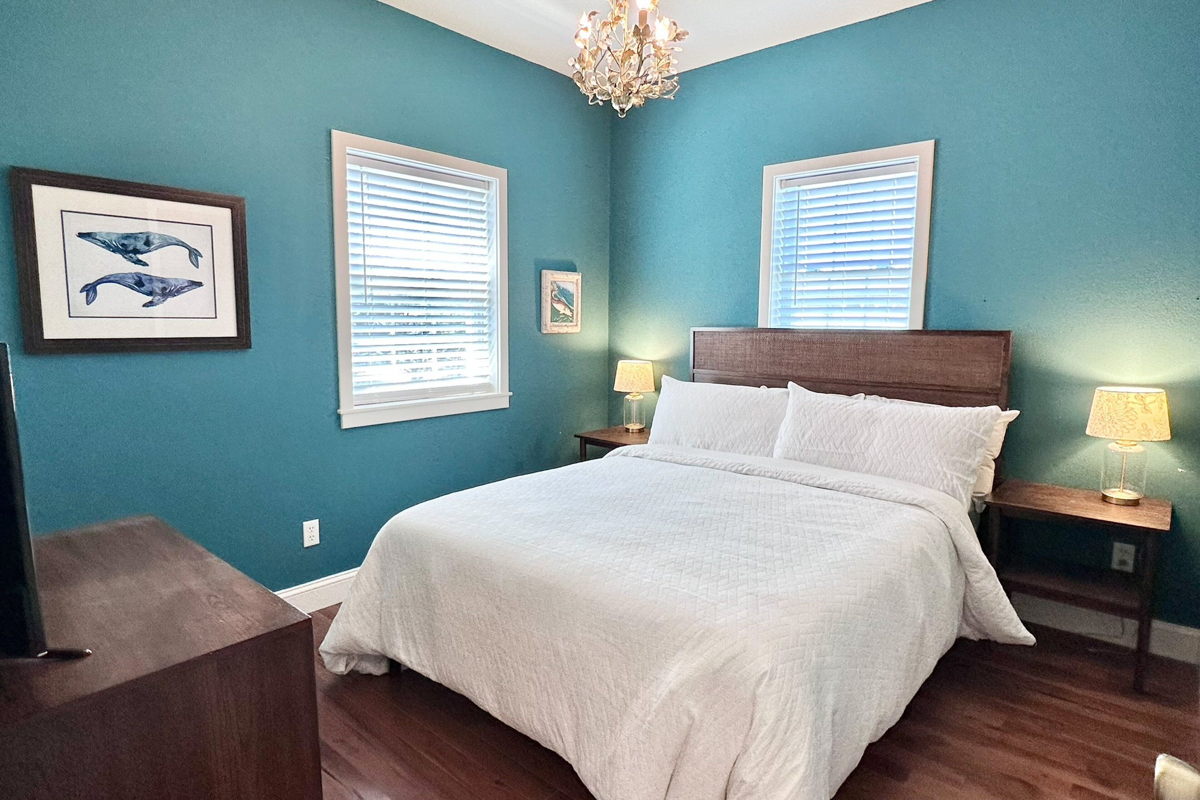 A bedroom with a queen-sized bed, two bedside tables with lamps, a chandelier, and two windows with blinds.