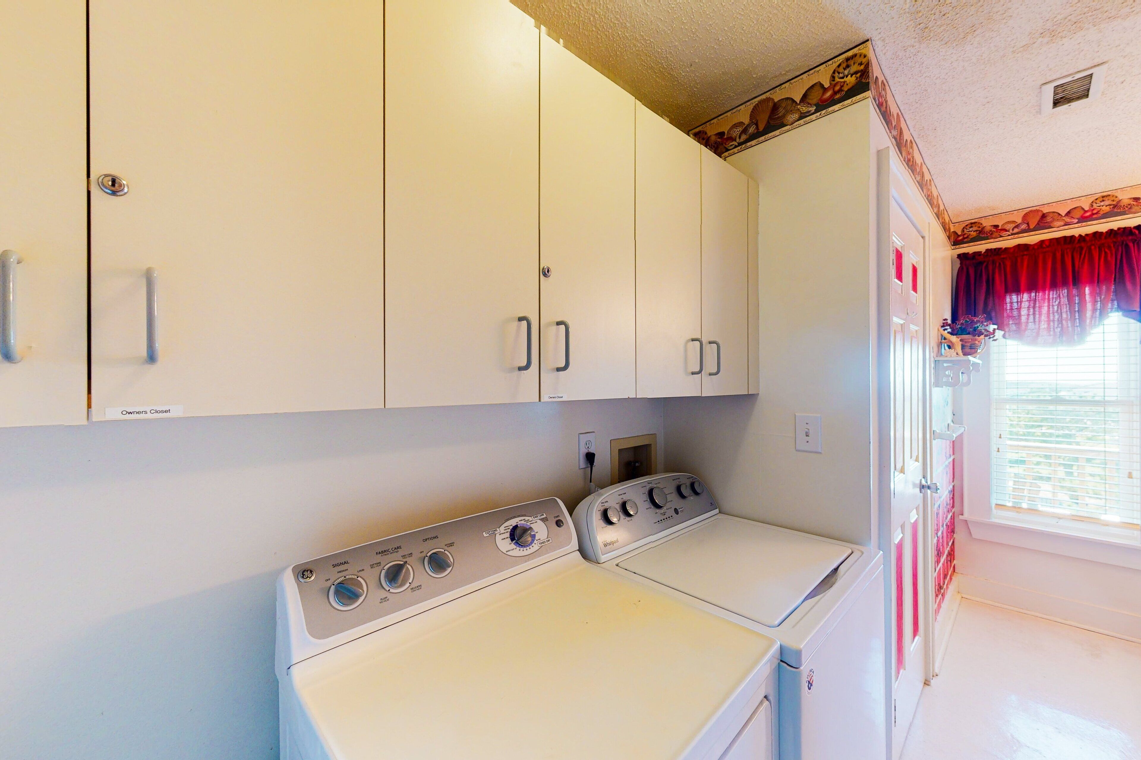 A laundry room with a white washing machine and dryer, white cabinets, and a window with red curtains.