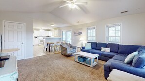 Uma sala de estar espaçosa com um sofá azul, uma poltrona estampada e uma mesa de centro. Há um ventilador de teto e uma cozinha ao fundo.